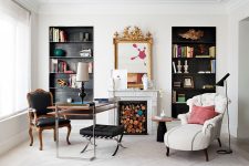 a fantastic home office with built-in shelves in black niches, a two tiered desk, a black chair and stool and a white lounger plus a fireplace with some wood