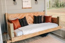 a farmhouse porch with a hanging bed of stained wood with printed pillows, sconces, an artwork and a rug is a very cozy space