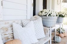 a farmhouse porch with a shabby whitewashed bench with pillows, potted blooms and some pumpkins is a cozy space