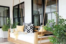 a front porch with an outdoor hanging bed with bright pillows and a blanket, with potted plants and a jute rug is a stylish space