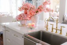 a glam white kitchen with shaker cabinets, gold handles, a gold pendant lamp and fixutres paired up with a chromatic sink