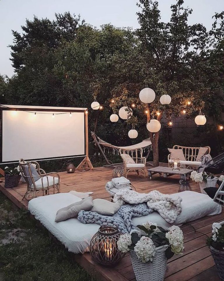 a gorgeous boho outdoor moovie space with rattan furniture, some mattresses, a movie screen and paper lamps plus lights
