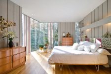 a light-filled mid-century modern bedroom with a glazed wall, stained furniture, a hairpin leg bed, potted plants and blooms
