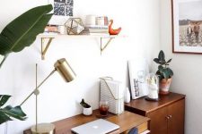 a little mid-century modern home office nook with a stained desk, a storage cabinet, a navy chair, a shelf, a gold lamp and potted plants