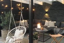 a lovely modern patio with a touch of boho, with wicker and metal furniture, a hanging macrame chair, potted greenery, candle lanterns and firewood