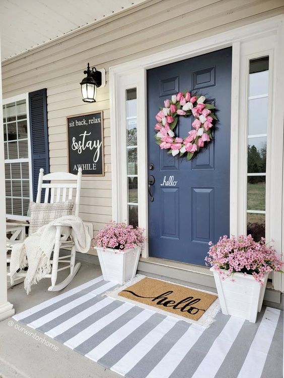 a lovely rustic porch styled for spring, with white furniture and plants, layered rugs and pink blooms plus a faux flower wreath