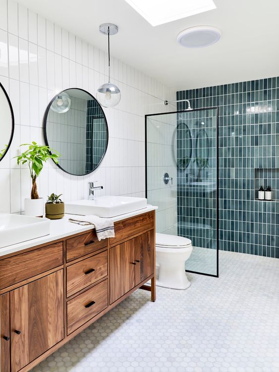 A mid century modern bathroom with blue skinny tiles in the shower, white hex tiles on the floor, a stained vanity and round mirrors