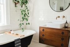 a mid-century modern bathroom with different white tiles, a black clawfoot tub, a floating vanity, potted greenery and a round mirror