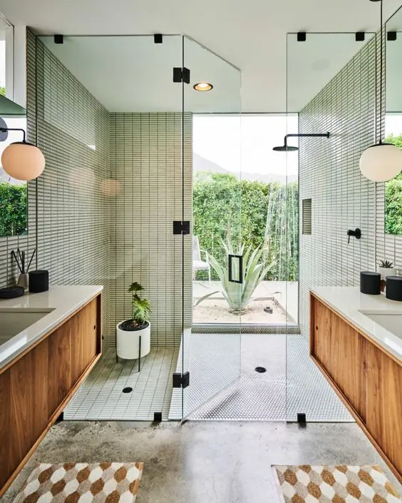 A mid century modern bathroom with skinny green tiles in the shower and a window, stained vanities, printed rugs and potted plants