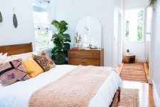 a mid-century modern farmhouse bedroom with stained furniture, neutral and printed bedding, potted greenery, a rounded mirror and a printed rug