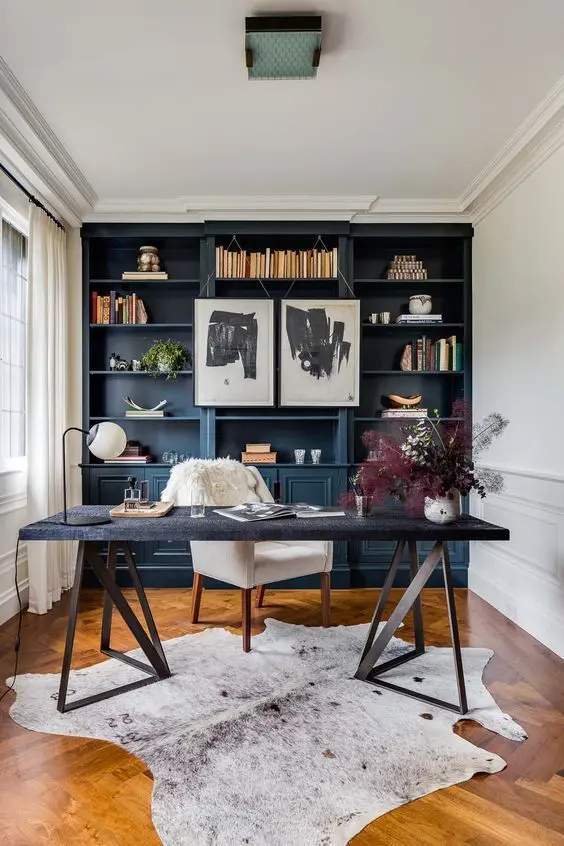 a mid-century modern home office with a navy built-in storage unit with shelves, a black trestle desk, a creamy chair and some leaves