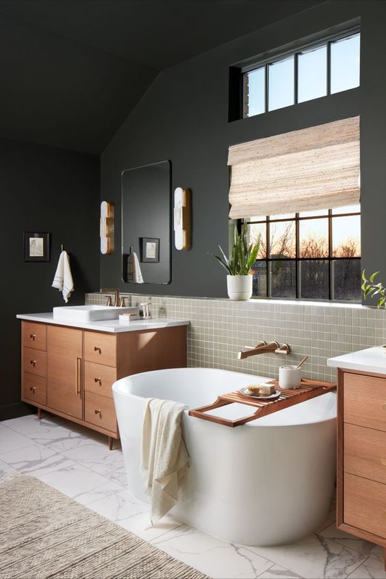 a moody mid-century modern bathroom with black walls, greige tiles and marble ones, an oval tub, stained vanities and potted plants