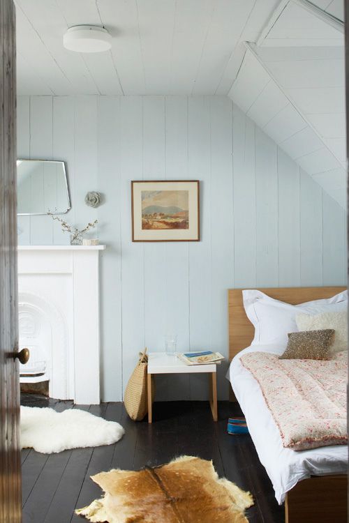 a pastel bedroom with light blue planked walls, a built-in fireplace, stained furniture, animal skin rugs, some art and a mirror