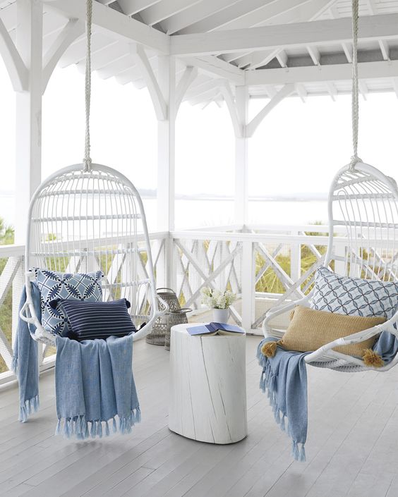 a pretty coastal porch with white hanging rattan chairs, a white side table, blue printed textiles and some blooms is a dreamy space to enjoy