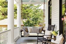 a pretty farmhouse porch with a striped rug, black rockers and a black hanging bench, printed pillows and greenery and blooms