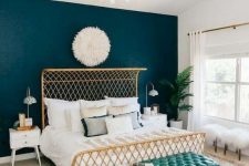 a pretty mid-century modern bedroom with a gilded bed, neutral and printed bedding, a gold chandelier, a turquoise bench, a printed rug and a potted plant