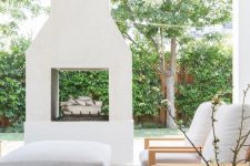 a pretty modern outdoor space clad with tiles, with a two-sided white fireplace, simple chairs and a low ottoman plus a lovely view of the garden