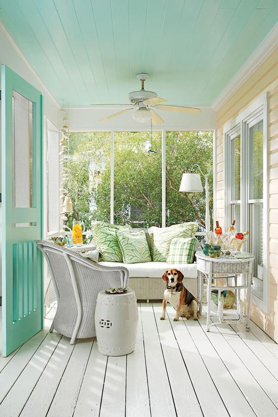 a pretty small coastal porch with a turquoise ceiling and doors, white wicker furniture, colorful pillows, side tables and some plants