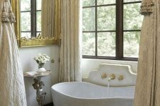 a refined Provence bathroom with a dark stained French window, a stone tub, neutral stone on the floor and neutral textiles and gold touches