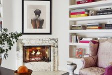 a refined home office with a marble clad fireplace, built-in shelves, a dark stained desk, a pink chair and some art and bright books
