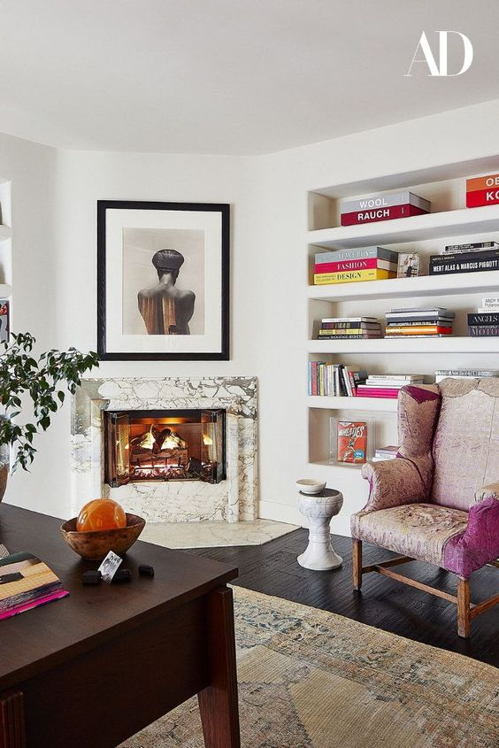 a refined home office with a marble clad fireplace, built in shelves, a dark stained desk, a pink chair and some art and bright books