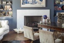 a refined home office with blue walls, built-in shelves, a fireplace, a vintage dark stained desk, grey chairs and a creamy sofa plus art