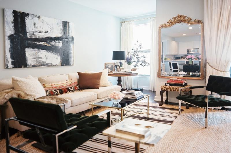 a refined living room with a neutral sofa, a dark green chair, a glass table, an oversized mirror in a gold ornated frame, chairs with chrome legs and armrests