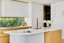 a refined mid-century modern kitchen with white and light-stained cabinets, a window backsplash, a sculptural kitchen island and gold touches