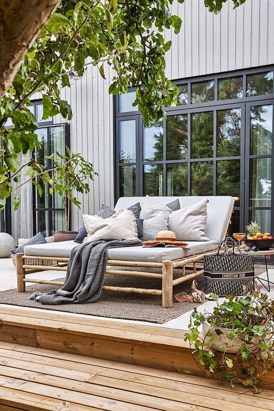 a relaxed deck with a large bamboo daybed with muted pillows and blankets, a side table, trees and greenery and vases