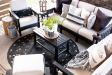 a rustic outdoor space wiht a stone clad fireplace, black furniture with white upholstery, a black table and a black jute rug, a chandelier is cool