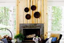a rustic porch with a stone fireplace and a wood cover, simple dark wicker furniture, decorative plates and printed and neutral textiles
