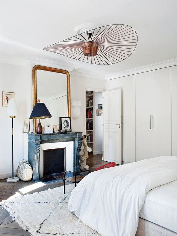 a serene Parisian bedroom with built in wardrobes, a fireplace with a blue mantel, a large mirror in a gilded frame and a bed with neutral bedding