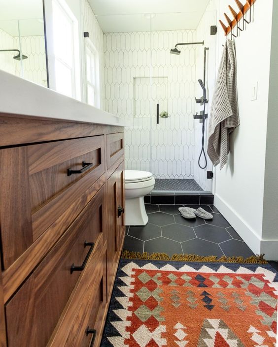a small and cozy mid-century modern bathroom with hex tiles on the floor, a boho rug, a stained vanity, black fixtures is chic