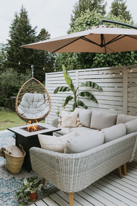 a small and very welcoming space with a white wicker corner sofa, a fire pit, a rattan chair and an umbrella plus a printed rug