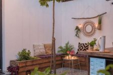 a small and welcoming outdoor space with a gravel floor, a pallet wooden bench, printed pillows and a rug, potted plants and greenery