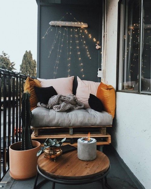a small modern balcony with a black accent furniture, a pallet loveseat, a round table, a potted cactus, string lights and greenery