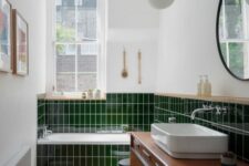 a small modern bathroom with bold grene tiles, a tub clad with them, a stained vanity, a sink and a round mirror and pendant lamps