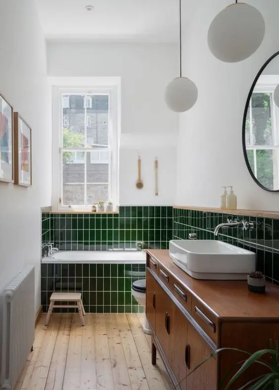 a small modern bathroom with bold grene tiles, a tub clad with them, a stained vanity, a sink and a round mirror and pendant lamps