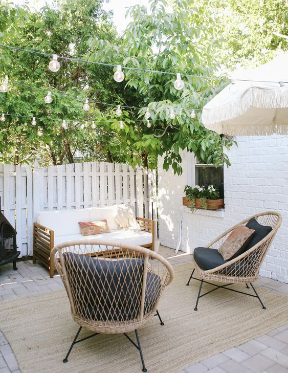 a small outdoor space wiht a wooden sofa, rattan chairs with black upholstery, greenery and string lights over the space