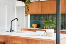 a stylish mid-century modern kitchen with light stained cabinets, white stone countertops, a green skinny tile backsplash, black fixtures and potted plants