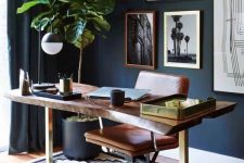 a stylish mid-century modern living room with navy walls, a rich-stained living edge desk, an amber leather chair, a potted tree and a gallery wall