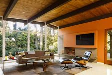 a super bright mid-century modern living room with a planked wood ceiling, an orange accent wall, a fireplace, a taupe sofa and a black leather chair