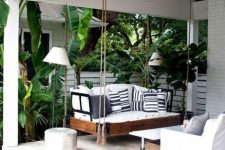 a tropical terrace with a hanging daybed with printed pillows, a coffee table and a white chair plus lots of tropical greenery around