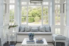 a vintage coastal interior in light blue and white, with lots of French windows and much light thanks to them, refined white furniutre, a chandelier and printed pillows