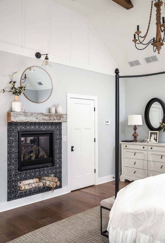 a vintage farmhouse bedroom with a black metal canopy bed, a built in fireplace with black tiles, a round mirror, a white dresser and a wooden chandelier