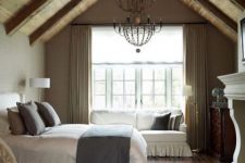 a vintage rustic bedroom with a wodoen slanted ceiling, a vintage fireplace, a bed with grey and white bedding, a sofa, a vintage chair and vintage chandeliers