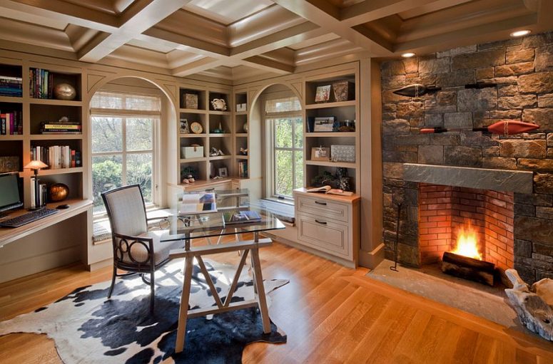 a welcoming and cozy home office with a stone clad fireplace, built-in shelves and niche shelves, a glass desk and a white chair