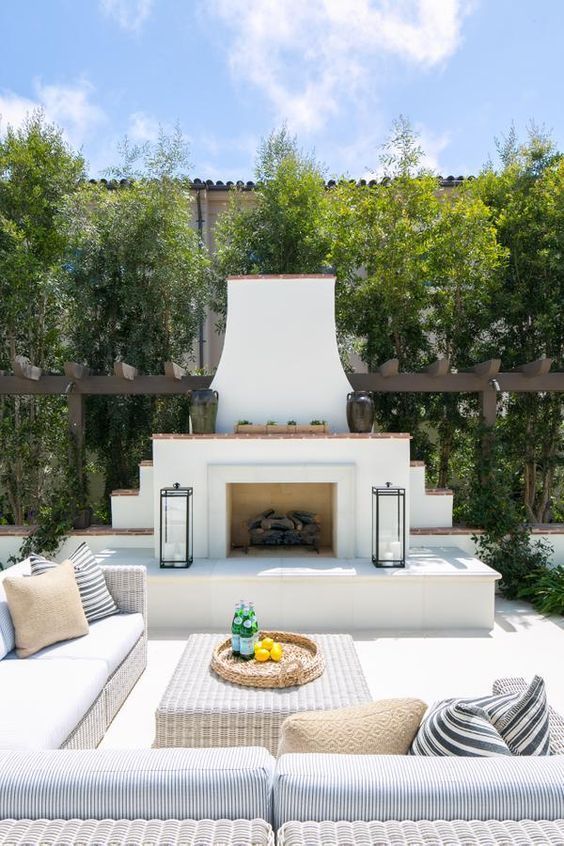 a welcoming coastal terrace with a white stucco fireplace, a white wicker sofa and an ottoman, candle lanterns and lots of greenery
