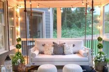 a welcoming farmhouse porch with a hanging sofa, white ottomans, a graphic rug, potted greenery and candle lanterns