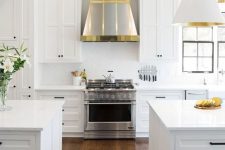 a white farmhouse kitchen with shaker style cabinets, black handles, silver hood with gold touches, white and gold lamps and chromatic appliances
